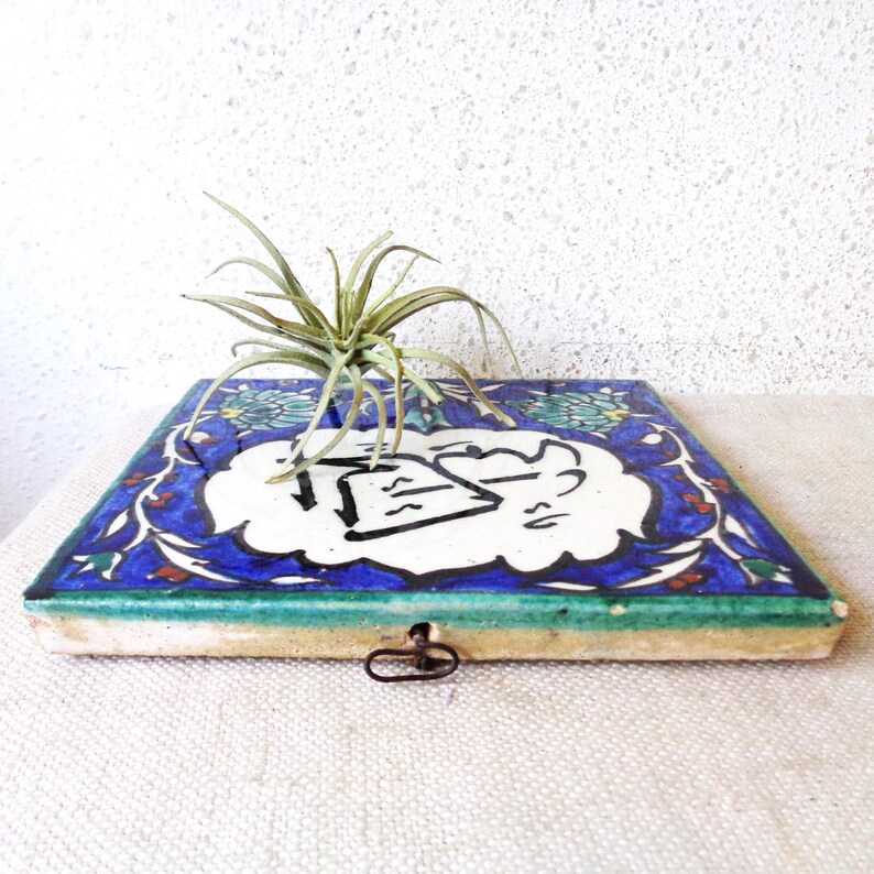 May include: A square, ceramic tile with a blue and white floral design, featuring a central white cloud-shaped element with black calligraphy. A small air plant sits atop the tile. The tile has a green border and a metal clasp.