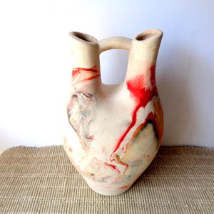 May include: A double-spouted ceramic vessel with a matte off-white base color. The surface features abstract, fluid swirls of vibrant red, charcoal gray, and muted ochre paint, creating a marbled effect across the rounded body and two necks.