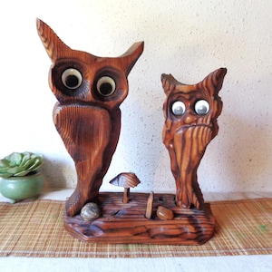 May include: Two wooden owl figurines with large, round eyes and a carved wood grain texture. The owls are mounted on a wooden base with a small mushroom and pebbles. The wood has a warm, brown tone, and the eyes are white with black pupils.