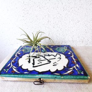 May include: Square, hand-painted ceramic tile with a blue floral border and a white center featuring black calligraphy. A small, green air plant sits atop the tile. The tile has a green edge and a metal clasp.