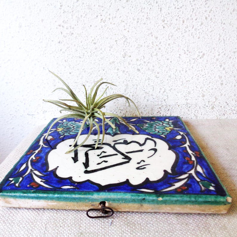 May include: Square, hand-painted ceramic tile with a blue floral border and a white center featuring black calligraphy. A small, green air plant sits atop the tile. The tile has a green edge and a metal clasp.