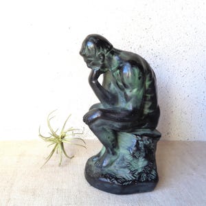 May include: A bronze-colored sculpture of a seated figure, with a hand resting on the chin, in a pose of contemplation. The statue has a green patina and sits on a stone base. A small, spiky plant is to the left of the statue.