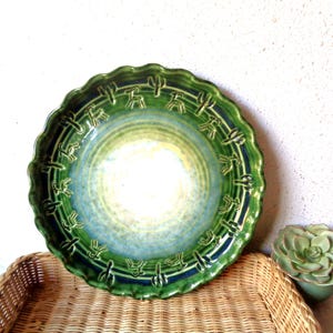 May include: A green ceramic plate with a scalloped edge and a cactus design. The plate has a gradient of colors, transitioning from green to blue and yellow in the center. The plate is sitting on a wicker basket.