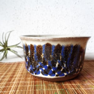 May include: A ceramic bowl with a wide, flared rim and a rounded base. The exterior features a decorative pattern of vertical, textured dark brown and vibrant blue brushstrokes over a cream-colored background, with a dark brown base.