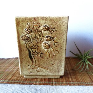 May include: A rectangular ceramic vase with a textured, speckled beige finish. The front features a raised, gold-colored tree design with textured, gold-colored fruit. The vase has a square base and is suitable for displaying flowers or as a decorative item.