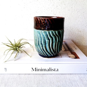 May include: A handcrafted ceramic mug with a textured, ridged design. The top portion is a dark brown, while the lower section is a teal blue. The mug is sitting on a white book with the word "Minimalista" printed on it.