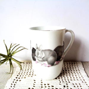 May include: White ceramic mug with a sleeping bunny illustration in shades of gray and pink. The mug has a gold rim and a curved handle. The design features a detailed depiction of a rabbit resting, adding a touch of whimsy to the drinkware.
