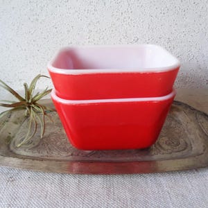 May include: Two square, red and white vintage Pyrex bowls stacked on top of each other. The bowls have a glossy finish and a white interior. They are sitting on a silver tray with an ornate design.