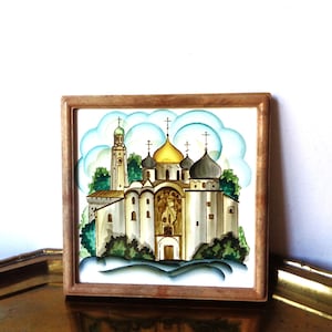May include: A square wooden-framed tile featuring a hand-painted illustration of an Orthodox church with a central gold dome, smaller grey domes, and a tall bell tower. The scene includes stylized green trees and blue clouds against a white background.