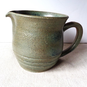 May include: A handmade stoneware pitcher with a curved spout and a rounded handle. The pitcher has a speckled, earthy green and brown glaze with subtle blue undertones. The body features horizontal grooves near the base, adding texture.