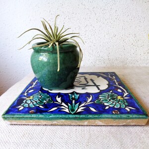 May include: A small, green ceramic pot with a plant sits on a square, hand-painted tile. The tile features a blue background with floral designs in teal, white, and yellow. The pot has a crackle glaze.