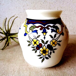 May include: A white ceramic vase with a rounded body and a wide opening. The surface features a decorative pattern of yellow flowers with green leaves, accented by dark blue, red, and yellow geometric lines encircling the upper section.