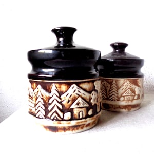 May include: Two ceramic jars with lids, featuring a brown and white mountain scene with trees and houses. The jars have a glossy brown glaze on the top and a matte brown glaze on the bottom.