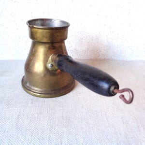 May include: A vintage brass coffee pot with a dark wooden handle and a curved metal hook. The pot has a wide base that tapers to a narrower neck and a flared rim. The handle is attached with a small metal plate and rivets. The pot is likely used for making Turkish coffee.