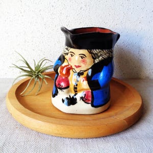 May include: A vintage ceramic pitcher featuring a painted figure in a black hat and blue coat. The pitcher has a black rim and a cream-colored base. It is placed on a round wooden tray with a small plant.