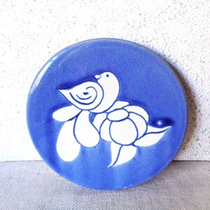 May include: A round, blue ceramic coaster with a white bird and floral design. The bird is perched above a stylized flower, creating a simple, elegant aesthetic. The coaster is a solid blue color with a glossy finish.