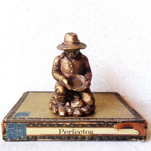 May include: A bronze-colored figurine of a prospector kneeling, holding a pan, atop a decorative box. The box is rectangular with gold and brown accents, and the word "Perfectos" is printed on the side.