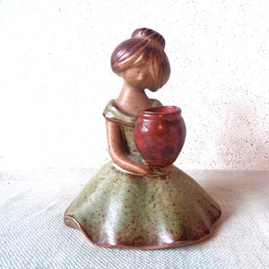 May include: A ceramic figurine of a woman in a dress, holding a small red vase. The dress is a muted green with brown speckles, and the figure has brown hair. The figurine is a decorative piece.