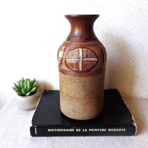May include: A vintage brown ceramic vase with a textured surface and a decorative design. The vase has a narrow neck and a wider base, with a cross-like pattern in the center. The vase is approximately 10 inches tall.