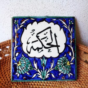 May include: Square ceramic tile with Arabic calligraphy in black on a white background. The tile has a blue border with floral and leaf designs in green, white, and red. The tile is placed on a woven wicker surface.