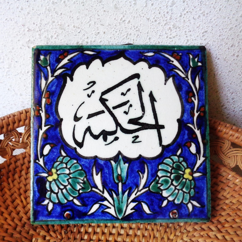 May include: Square ceramic tile with Arabic calligraphy in black on a white background. The tile has a blue border with floral and leaf designs in green, white, and red. The tile is placed on a woven wicker surface.