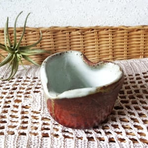 May include: A heart-shaped ceramic bowl with a rustic, handmade appearance. The bowl features a reddish-brown exterior, a white rim, and a pale blue interior. The bowl is placed on a white crocheted surface, with a woven basket in the background.