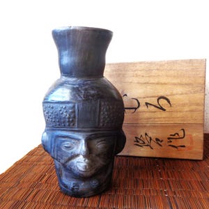 May include: A dark blue ceramic vase with a face design, featuring a detailed head and a flared top. The vase is placed on a woven mat, with a wooden box in the background.