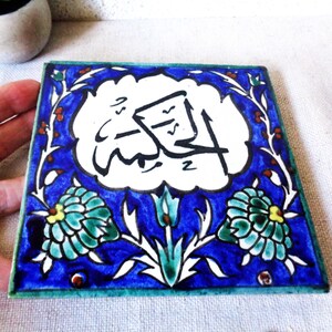 May include: Square ceramic tile with a blue border and a central design featuring Arabic calligraphy in black on a white background. The tile is decorated with floral motifs in shades of blue, green, and yellow.