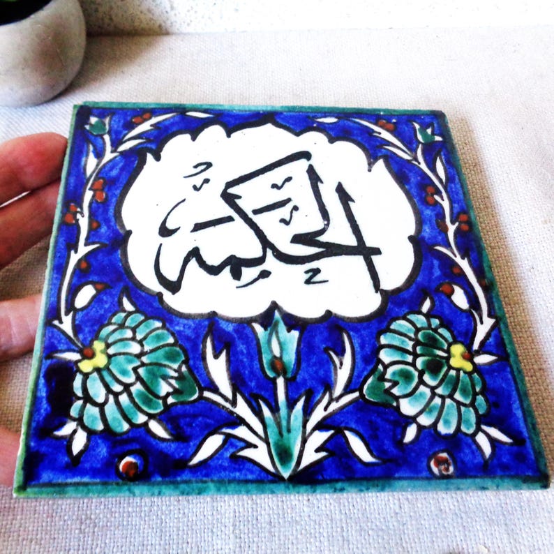 May include: Square ceramic tile with a blue border and a central design featuring Arabic calligraphy in black on a white background. The tile is decorated with floral motifs in shades of blue, green, and yellow.