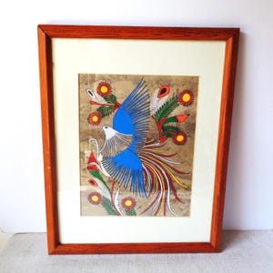 May include: Framed artwork featuring a vibrant bird with blue wings and a white head, set against a textured background. The bird is surrounded by colorful floral and leaf accents in red, yellow, and green. The artwork is encased in a wooden frame.