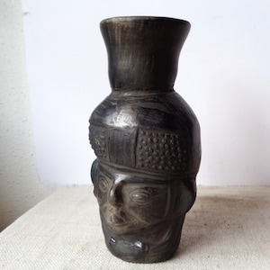 May include: A dark charcoal-colored ceramic vessel shaped like a human head. The object features a wide, flared neck opening, a textured band across the forehead with small raised dots, and stylized facial features including eyes and a mouth.