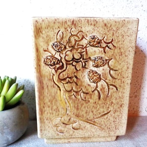 May include: A rectangular, beige ceramic vase with a textured surface. The vase features a raised, stylized floral design with gold glitter accents. The vase is a decorative item.