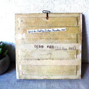 May include: Back of a vintage tile with a metal hanger. The tile is beige with horizontal rectangular indentations and white tape. Handwritten text reads "Bot at the Dead Sea, Indian Jerusalem 1965" and "DEAD SEA (Bet Haare 1965)".