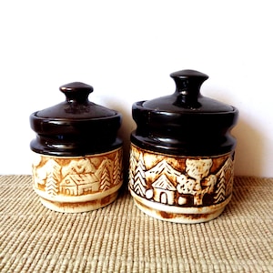 May include: Two ceramic canisters with dark brown lids and cream-colored bases. The lower sections feature embossed landscape designs of houses, trees, and mountains with brown detailing. Each container has a matching removable lid with a knob.