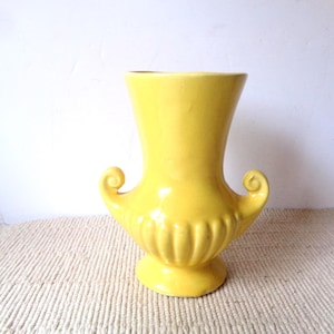 May include: A bright yellow ceramic vase featuring a flared rim, a wide neck, and a bulbous base with vertical fluted detailing. The vase includes two symmetrical scroll-shaped handles on either side and rests on a circular pedestal foot.