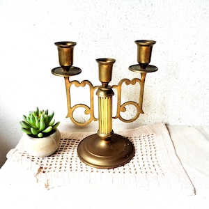 May include: A vintage brass candelabra with three candle holders. The candelabra has a round base, a central column with vertical ridges, and decorative arms. A small succulent plant in a white pot sits to the left of the candelabra.