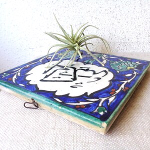 May include: Square ceramic tile with a blue and white floral design, featuring a central white emblem with black calligraphy. The tile has a green border and a small metal loop. A small air plant sits atop the tile.