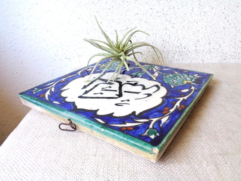 May include: Square ceramic tile with a blue and white floral design, featuring a central white emblem with black calligraphy. The tile has a green border and a small metal loop. A small air plant sits atop the tile.
