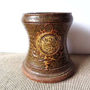 May include: A brown ceramic beer stein with a decorative lion emblem. The stein has a wide base and a narrower middle section, with a textured surface and a dark brown glaze. The emblem is in a lighter brown color.