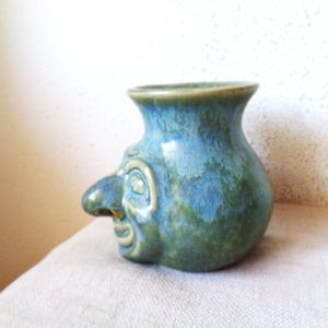 May include: A ceramic vase with a face sculpted on the side. The vase is glazed in shades of blue and green, with a prominent nose and detailed facial features. The vase has a rounded body and a flared rim.