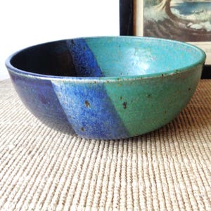 May include: A round ceramic bowl featuring a multi-colored glaze with distinct sections of deep navy blue, vibrant cobalt blue, and a speckled teal green. The surface has a smooth, glossy finish with subtle variations in color and texture throughout.