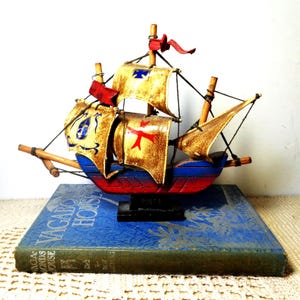May include: A model ship named Pinta with a red and blue hull, featuring tan sails decorated with red and blue cross emblems. The ship has wooden masts, black rigging, and a small red flag at the top, all mounted on a black base.