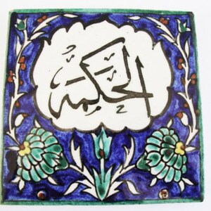May include: Square ceramic tile with a white center featuring black Arabic calligraphy. The tile has a blue border with floral and leaf designs in green, white, and orange. The edges are trimmed in green.