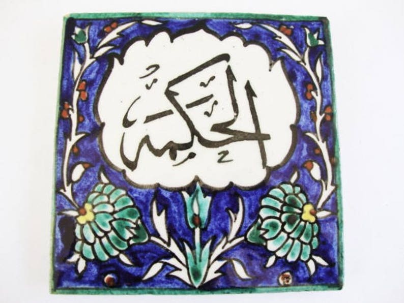May include: Square ceramic tile with a white center featuring black Arabic calligraphy. The tile has a blue border with floral and leaf designs in green, white, and orange. The edges are trimmed in green.