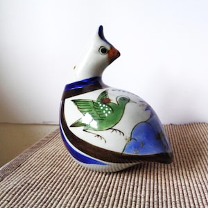 May include: A ceramic bird figurine with a white body, blue and brown accents, and a green bird illustration. The bird has a long neck and a pointed beak. The figurine is on a woven surface.