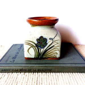 May include: A square ceramic vase with a speckled off-white body and a wide, rounded rim in a deep orange-brown glaze. The front features a hand-painted design of a dark blue flower with green leaves and stems, accented by a thin black line.