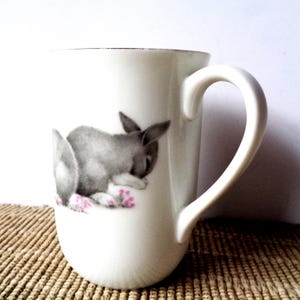 May include: White ceramic mug featuring a gray and white illustration of a sleeping bunny with pink flowers. The mug has a curved handle and a gold rim. The mug is sitting on a woven surface.