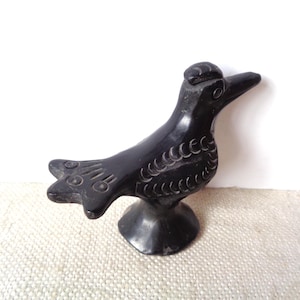 May include: A black, carved bird figurine with a detailed design. The bird has a long beak, a small crest, and a detailed tail. The figurine is mounted on a round base and is set against a neutral background.