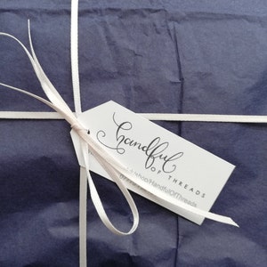K&ouml;nnte beinhalten: Ein verpacktes Geschenk mit marineblauem Seidenpapier, gebunden mit wei&szlig;em Band. Ein wei&szlig;es Etikett mit dem Schriftzug "Handful of Threads" in eleganter Schrift ist mit einem hellrosa Band befestigt.