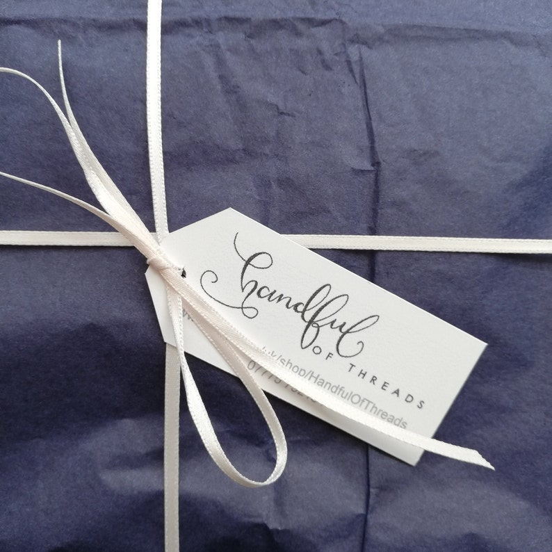 K&ouml;nnte beinhalten: Ein verpacktes Geschenk mit marineblauem Seidenpapier, gebunden mit wei&szlig;em Band. Ein wei&szlig;es Etikett mit dem Schriftzug "Handful of Threads" in eleganter Schrift ist mit einem hellrosa Band befestigt.